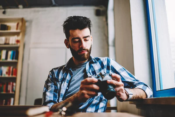 Pensive bearded young man holding vintage camera in hands and viewing photos on device sitting in college.Professional photographer making setting on gadget working in studio