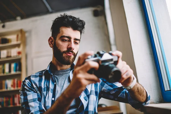 沉思的专业摄影师拿着老式设备 看着坐在自己的工作室的照片 胡子时髦的家伙穿着休闲装观看照片在相机和设置 — 图库照片