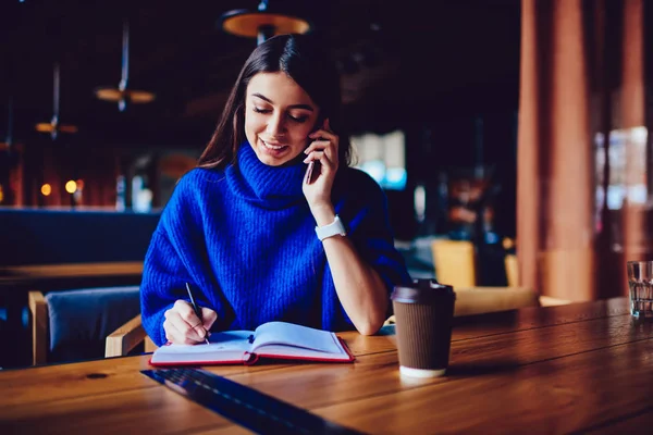 キャンパスカフェで勉強中に友人に電話をかける携帯電話を持つ陽気な女性 メモを書いたりカフェに座っている間に電話で話す4Gインターネットに接続された笑顔のヒップスターの女の子 — ストック写真