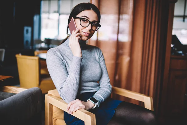 Smiling hipster girl calling customer service for consulting using mobile phone indoors while looking aside, positive woman in trendy spectacles having cellphone conversation in break of work