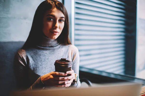 コワーキングスペースでノートパソコンでコーヒーブレイクを楽しむカメラを見ている魅力的な女子学生 カフェテリアで休みながらカップでポーズをとる白人美しいブルネットの女性 — ストック写真