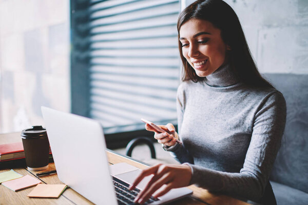 Весёлая женщина-фрилансер устанавливает приложение на современном ноутбуке, держа современный смартфон в помещении, успешная улыбающаяся женщина проверяет уведомления сидя в кафе
