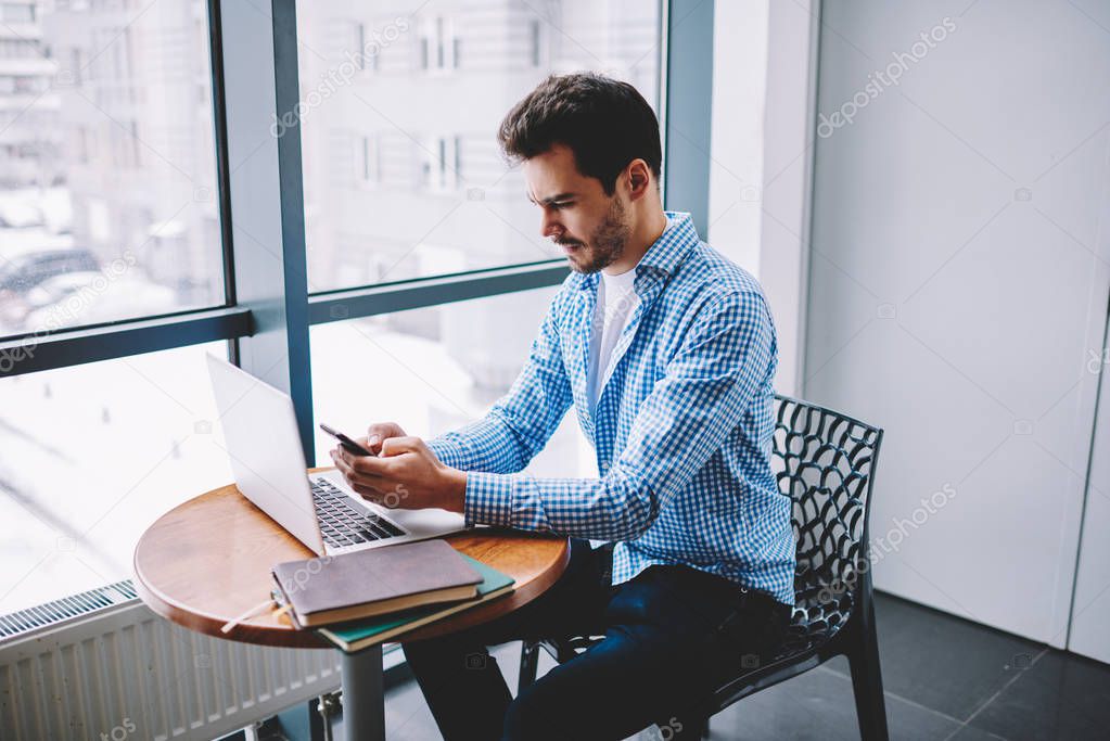 Serious man feeling puzzled on received email from work connected to internet on cellular for making communication online, concentrated male freelancer sharing information via smartphone and laptop