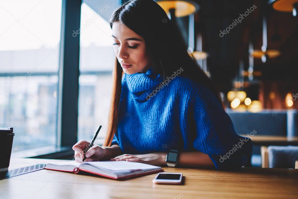 Serious thoughtful brunette hipster girl preparing for exam during resting at cafeteria, pondering female student learning at university campus while modern mobile phone staying at wooden table