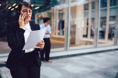 Elinde kağıt muhasebe raporları ile ofis binası yakınında açık havada dururken mobil konuşma sırasında finansal bilgileri tartışırken zarif kıyafet giymiş emin kadın bankacı