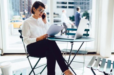 Pozitif kadın tüccar reklam alanında kağıt belgeler ile uzaktan çalışırken iş web sitesinde finansal haber okuma, masada oturan optik gözlük başarılı girişimci