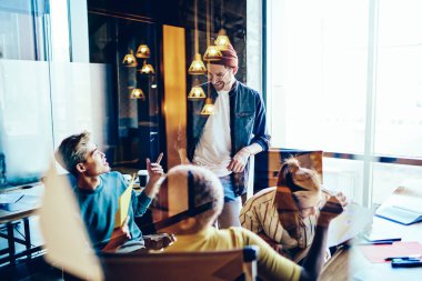 Gündelik giysiler giymiş pozitif gençler modern ofiste, cam duvarların arkasında beyin fırtınası yaparken eğleniyorlar. Çalışırken gülen başarılı hipster öğrenciler.