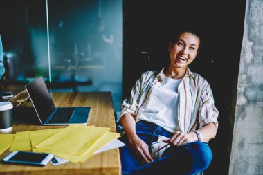 Modern ofis bilgisayarında modern dizüstü bilgisayarın başında gülen sıradan giyinmiş pozitif Japon genç bir kadın. Neşeli Çinli hippi kız netbook 'ta çalışırken mola verdi.