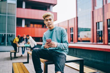 Sıradan giyinmiş Çinli bir gencin portresi kameraya bakarken açık havada oturan ve 4G internet bağlantısı kullanan modern akıllı telefon cihazına mesaj yazıyor. Japon hipster blogcu