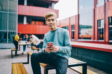 Açık havada oturan modern akıllı telefon cihazındaki uygulamayı güncellerken kameraya gülümseyen neşeli hipster blog yazarının portresi. Hücresel ağlarda sosyal ağlarda olumlu Japon genç adam sohbet ediyor.