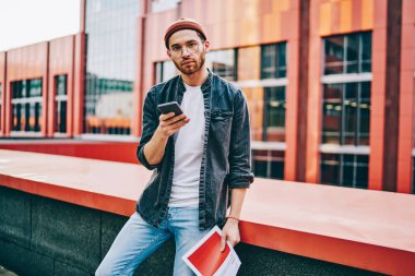 Modern akıllı telefon cihazını tutarken kameraya bakan ve sosyal ağlarda arkadaşlarıyla sohbet eden 20 yaşındaki sıradan giyinmiş bir hipsterin portresi şehir ortamında 4G internet bağlantısı kullanıyor.