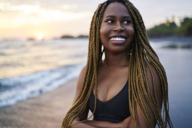 Sahilde sabah eğitimi öncesi dreads ile Neşeli afro amerikan koşucu, pozitif koyu tenli kadın aktif giyim sahil şeridinde ayakta giyinmiş ve yüzünde sevimli bir gülümseme ile uzağa bakıyor