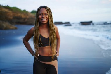Okyanus suyu ile yıkanmış plajda ayakta dreads ile pozitif neşeli sportwoman Portresi, sahilde kamera gülümseyerek egzersiz için aktif giysiler giymiş mutlu koyu tenli genç kadın
