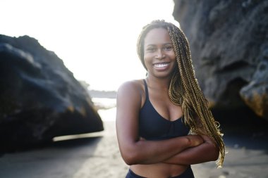 Neşeli afro amerikan genç kadın spor sevgilisi portre kamera önünde elleri çapraz iken gülümseyen, mutlu kadın arka planda kayalar ile açık havada ayakta egzersiz için aktif üst giymiş