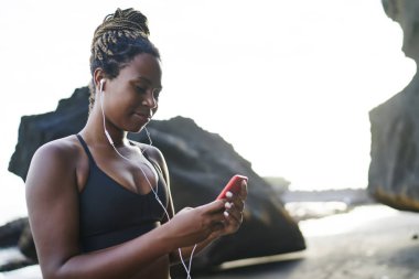 Kayalar yakınında dururken modern cep telefonu ve elektronik kulaklık uygulama kullanarak açık hava egzersiz için üst arama favori çalma listesi çekici afro amerikan sportif kadın