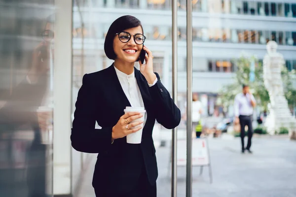 メガネをかけた陽気な女性トレーダーは おいしいコーヒーで屋外で時間を楽しみながら スマートフォンの会話中に良い金融ニュースを得ることに満足し ダウンタウンの企業の世界の概念 — ストック写真