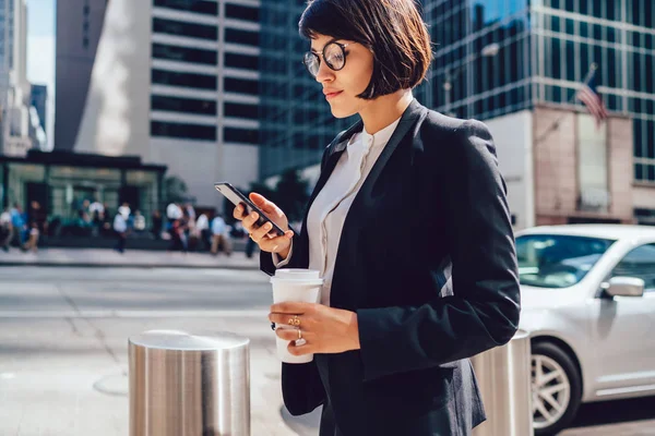 Young female director reading financial news on way to work using 4g connection on modern cellular phone, intelligent entrepreneur with coffee to go concentrated on received email from partner