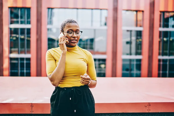 African american woman in eyeglasses explaining some information during phone conversation on modern smartphone using roaming connection in urban setting.Dark skinned female calling on cellular