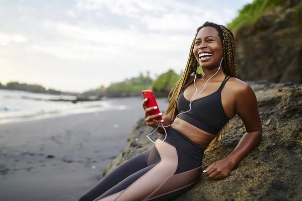 Happy afro american runner with dreads enjoying cool audio songs in earphones connected to smartphone resting on sea coast, positive dark skinned jogger in active clothes for training listening music