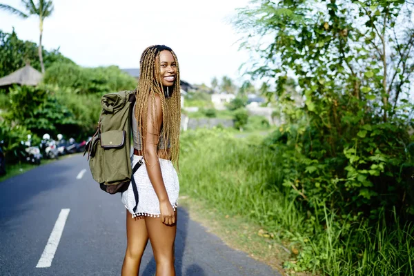 Portrait of happy afro american tourist with dreads smiling at camera during summer trip in tropical island, positive dark skinned traveller with backpack standing on road enjoying weekends