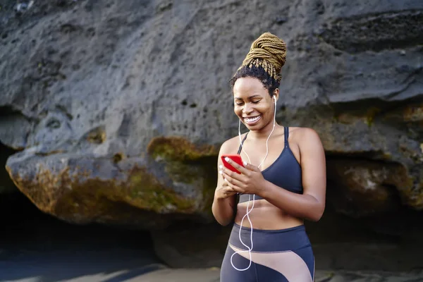 Happy dark skinned female jogger happy from cool music on radio for morning workout outdoors, cheerful excited woman listening audio from player in headphones connected to cellular before training