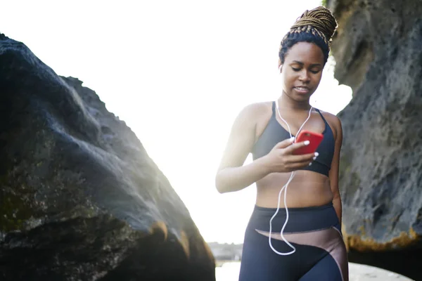 年轻女子穿着运动服 穿着运动服 穿着运动服的非洲裔美国女赛跑选手通过智能手机下载音乐歌曲 在岩石附近进行训练 — 图库照片