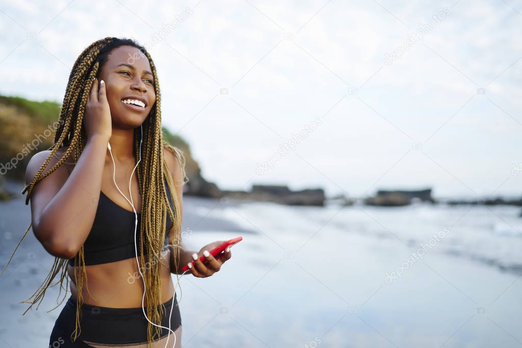 Positive smiling woman dressed in active wear laughing standing on sea coastline in morning time and listening music via earphones, happy dark skinned female jogger in sport clothes using cellular