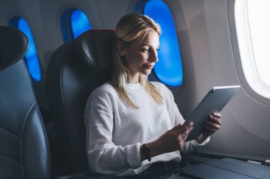 Genç beyaz kadın yolcu modern elektrokromik teknolojiyle geniş kontrollü pencerenin yanında geniş koltukta rahat bir uçuş yapıyor. Turist kadın gemide kablosuz bağlantı kullanıyor