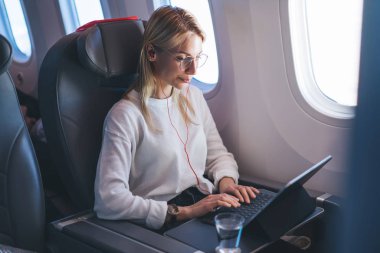 Beyaz bir kadın uçuş sırasında serbest çalışıyor ve kablosuz internet erişimini kullanıyor. Genç sarışın kadın müzik dinliyor, klavye e e-postası uçaktan iniyor.