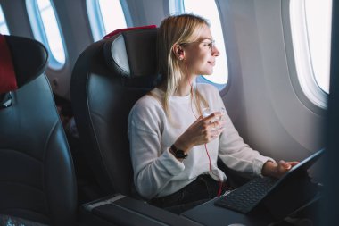 Genç beyaz kadın, kabinde oturmuş kulaklıkla müzik dinlerken ve su içerken rahat uçuşunun keyfini çıkarıyor. Kabinde internet erişimi, pencerenin yanında yolcu