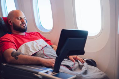 30 yaşında, beyaz bir adam birinci sınıf uçuş sırasında battaniyeyle sarılmış, geniş koltukta uyuyor. Modern eğlence sistemiyle rahat bir uçakta uyuyan erkek uçak yolcusu.
