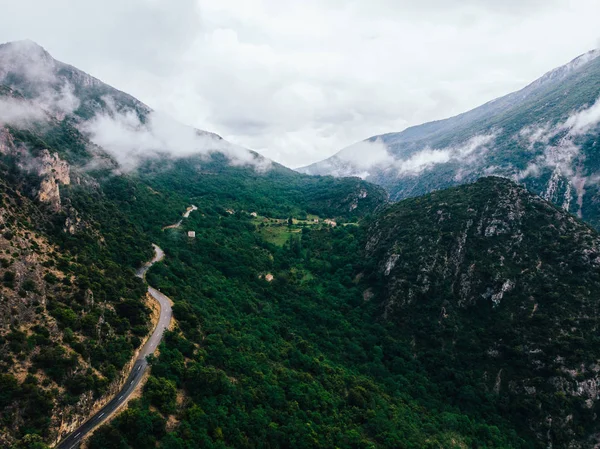 Aerial scenery view of asphalt road pass in rocky mountains with green vegetation covered with fog after rain. Bird\'s eye view of rocky hills with picturesque bike path above which hung heavy clouds
