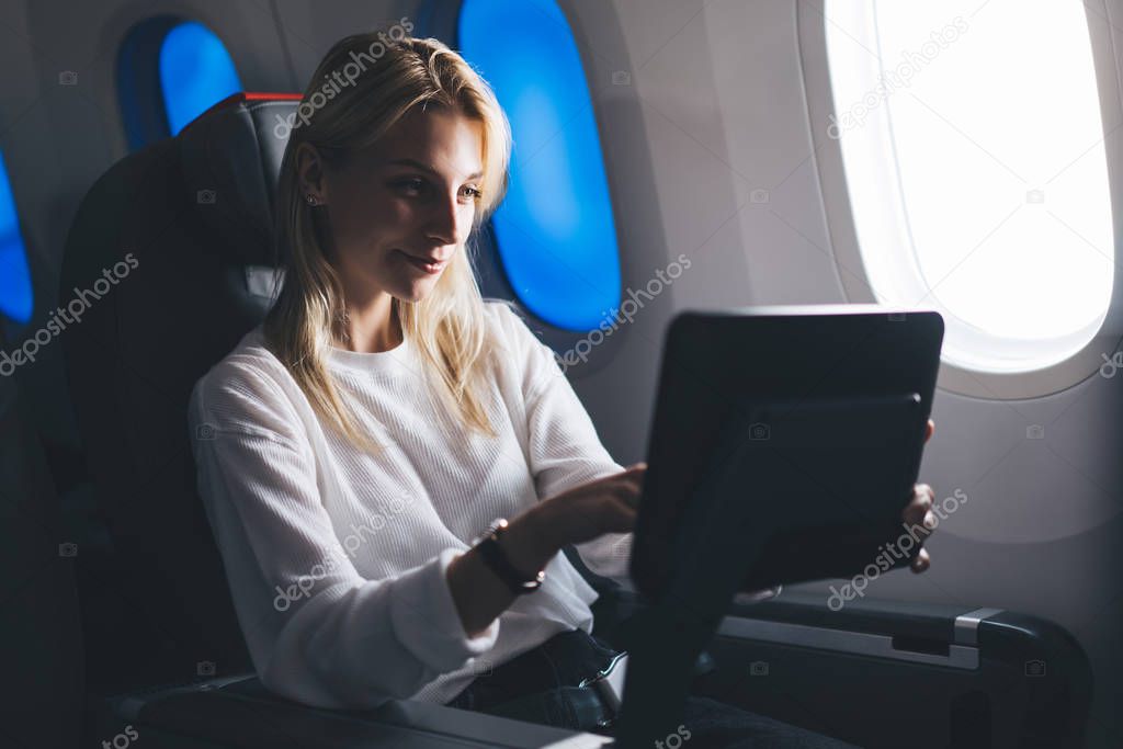 Caucasian hipster girl passenger touching with hand LCD screen monitor while choosing entertainment during comfortable flight.Playing games, shopping store,personal television.Movie selection on board
