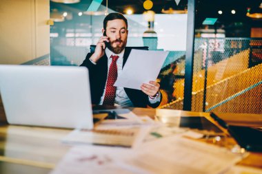 Resmi olarak sakallı deneyimli girişimci finansal raporları okuyarak ve veri taraması için akıllı telefondan yöneticiyi arayarak giyer. Ofiste cep telefonuyla konuşan kağıt belgeli bir işadamı