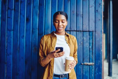 Neşeli Afro-Amerikalı genç bir kadın elinde kahve tutarak şehir ortamında duvara karşı duran umumi 4G interneti kullanarak cep telefonuyla sohbet ediyor. Pozitif koyu tenli hippi blog yazarı.