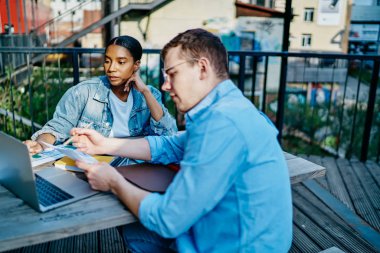 Yoğunlaşmış koyu tenli hippi öğrenci, modern dizüstü bilgisayarda beyaz meslektaşlarıyla birlikte ders izliyor. Tahta masada oturuyorlar ve seminer için hazırlanıyorlar.