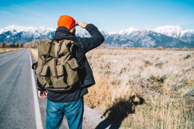 Yürüyüş için şık sırt çantasıyla asfalt yolda dikilirken iyi bir ritm ve tempoyla tepeye çıkmak için dağlara giden yolu arayan genç erkek yürüyüşçünün arka görüntüsü, nefes kesici kavram.
