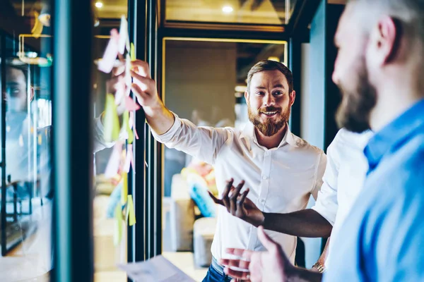 Cheerful bearded proud ceo in white shirt laughing while pointing on glass wall with colorful stickers and discussing solution with colleagues.Positive caucasian colleagues collaborating in office