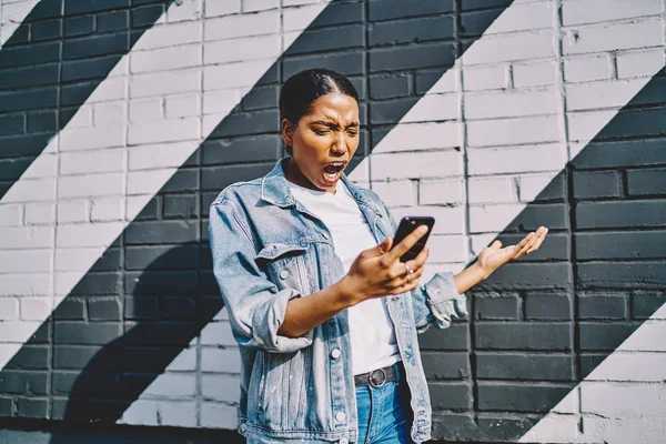 Angry african american young woman shocked with received notification with unhappy news standing in urban setting.Shocked dark skinned hipster girl has problem in mobile app on modern cellular