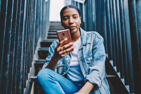 Güzel Afro-Amerikan genç bir kadının portresi kameraya bakıyor, elinde akıllı telefon cihazı var ve 4G internet bağlantısı üzerinden uygulama yüklüyor. Yakışıklı, koyu tenli hippi kız cep telefonuyla sohbet ediyor.