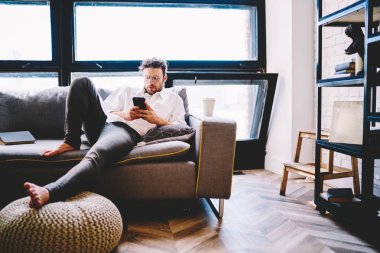 Gözlüklü, ciddi bir beyaz adam cep telefonu numarasıyla ev içi koltukta yatıyor, bin yıllık hipster adam akıllı telefondan posta ve mesajları kontrol ediyor ve online dağıtım numaralarını arıyor.