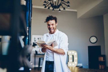Heyecanlı hipster adam komik edebiyat kitapları okuyor komik espriler yapıyor ve ev içinde gülüyor, olumlu gülümseyen adam şık bir dairede dururken elinde kitap tutuyor.