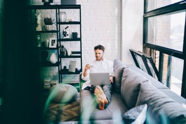 Modern tasarım oturma odasındaki koltukta dinlenen neşeli hippi adam dizüstü bilgisayar ve kablosuz internet üzerinden online alışveriş yapıyor. Tarayıcıda arama yapmak için kartvizitten bilgi tutan ve okuyan adam.