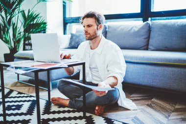 Yetenekli adam, modern apartmanın zemininde oturarak ve internet sitelerini taramak için 4G kablosuz internet kullanarak uzaktan çalışma ve başlangıç projesine odaklanmış genç hippi adam, netbook üzerinden kısa mesaj yazıyor.