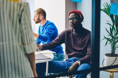 Fikirler ve iş akışı üzerine yoğunlaşan çok ırklı çalışanların kesilmiş görüntüsü. Masaüstünde oturan ve meslektaşlarının açıklamalarını dinleyen ciddi bir Afrikalı Amerikalı.