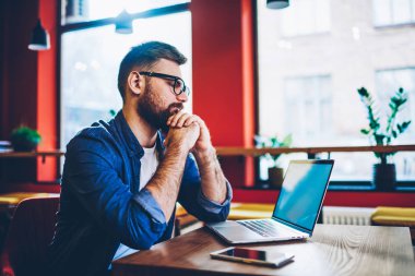 Ciddi bir erkek moda göz giyimi uzmanı içerik web sitesinde modern teknolojinin tebligatı hakkında haberleri okurken aynı zamanda kafenin içinde uzaktan çalışıyor ve ücretsiz internet bağlantısı kullanıyor.