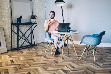 Hipster bir adamın ev içinde dizüstü bilgisayarla çalışırken kahve molasından keyif aldığını düşünmek şık tasarım, düşünceli erkek metin yazarı web sayfasında yeni bir makale fikri düşünüyor.