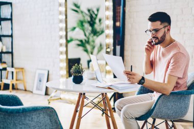 Optik gözlüklü, kağıt belgelerle uzaktan yakından ilgilenen, iş arkadaşına danışmanlık yapması için telefon eden, modern akıllı telefon cihazında 4G internet kullanan ve ev masasında oturan bir adam.