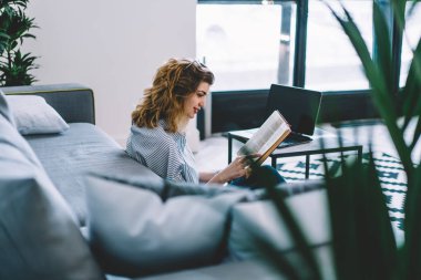 Modern dizüstü bilgisayarın üstünde oturmuş, şık iç dekorasyonlu bir şekilde kitap okuyan gülümseyen genç bir kadın. Evde boş zamanlarında edebiyat entrikalarından hoşlanan pozitif hipster kız.