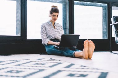 Gülümseyen genç bir kadın dizüstü bilgisayarda klavye kullanıyor ve uzaktan kumanda uygulamasını güncelliyor. Büyük pencereli, modern bir dairede yerde oturuyor. Hipster kızı netbook üzerine ders için metin yazıyor.